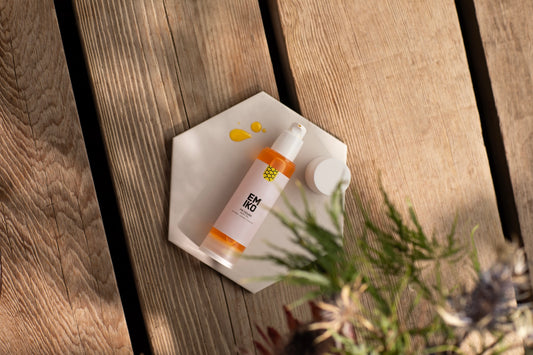 A bottle ofEmiko skincare Organic Body Oil to be use din the bath as well, botanicals, displayed on a small white hexagonal dish on a rustic wooden plank surface with blurred green foliage in the foreground.