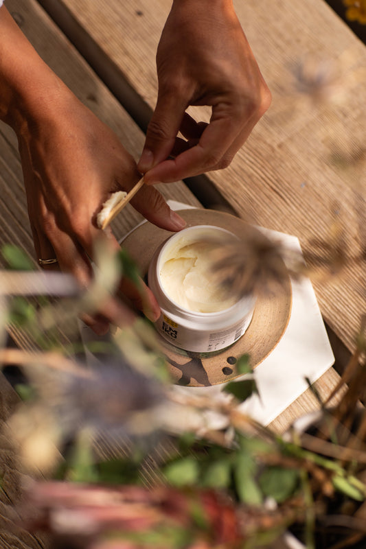 A close-up lifestyle shot of a person's hands applying Emiko Skincare & Relax Hand and Body Butter, from a white cosmetic jar onto a wooden spatula, set on a rustic wooden table with blurred natural elements in the foreground.
