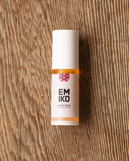 A bottle of EMIKO Bare Face Balancing Oil 30ml with a white cap and label, resting on a rustic wooden surface.