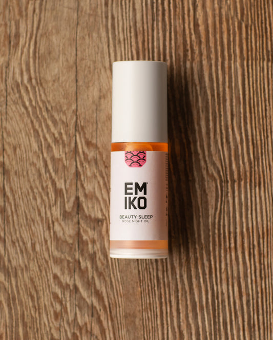 A bottle of EMIKO Beauty Sleep Rose Night Oil with a white cap and a white label featuring orange and pink accents, set against a rustic wooden background.