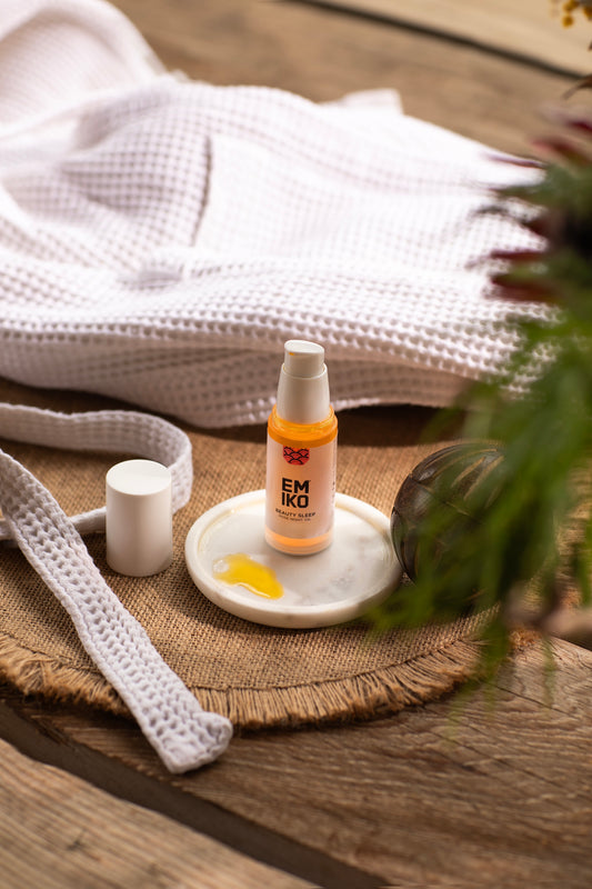30ml Beauty Sleep Rose Night Oil Emiko Skincare of orange-colored skincare oil with a white pump sits on a white dish on a woven mat, styled with a white waffle-knit towel, a crochet strap, and green leaves on a rustic wooden surface.