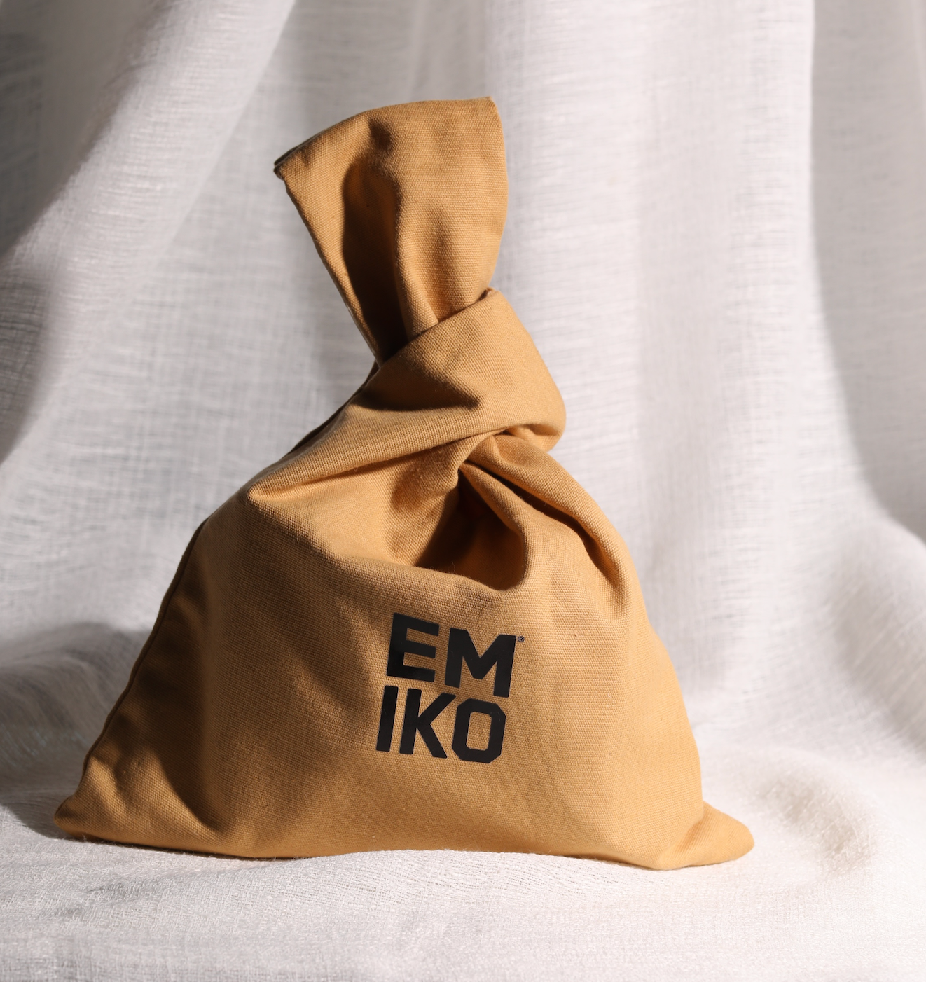 EMIKO SKINCARE 100% cotton Japanese knot bag, tan colour with the brand name 'EMIKO' printed in bold black letters, set against a draped white linen background.