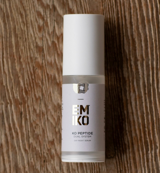 A bottle of EMKO KO Peptide Dual System Day Night Serum win an air pump dispenser white cap, resting on a brown wood grain background. It has advanced peptides for day and night use. 