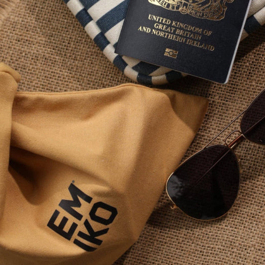 A tan Japanese 100% cotton knot bag with the brand name 'EMIKO' printed in black, styled on a burlap surface next to a navy and white striped lap top case, sunglasses, and a British passport, carrying 5 travel size products which is the EMIKO core ritual; Deep Clean, Nippon, Power, Boost, Glow.