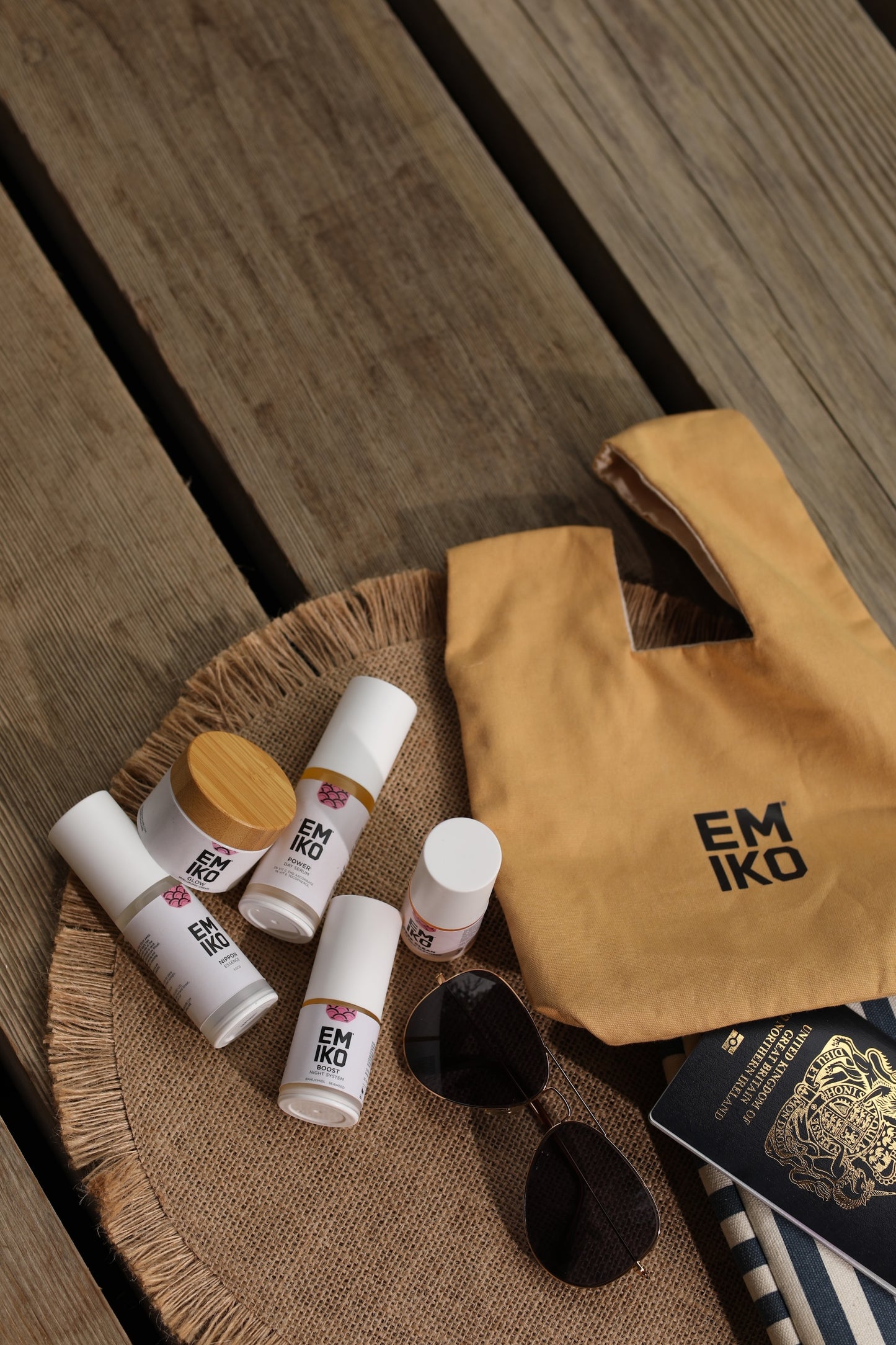A tan Japanese 100% cotton knot bag with the brand name 'EMIKO' printed in black, styled on a burlap surface next to a navy and white striped lap top case, sunglasses, and a British passport, carrying 5 travel size products which is the EMIKO core ritual; Deep Clean, Nippon, Power, Boost, Glow.