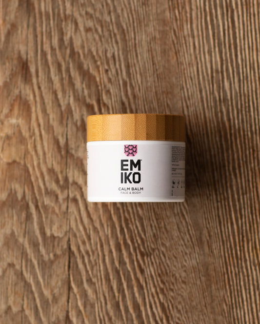 A 50ml white jar of EMIKO Calm Balm for sensitive reactive skin  with a light bamboo lid, placed on a rustic wooden surface.