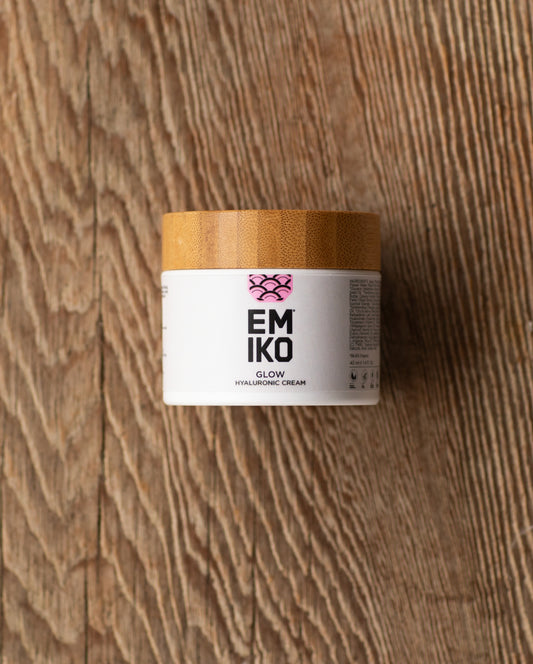 A white cosmetic jar with a light wood lid labeled EMIKO GLOW HYALURONIC CREAM' on a rustic wooden background.