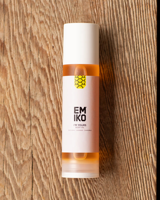 A bottle of Emiko Skincare I'm Yours Body Oil with a white cap and label, showing the amber-colored oil inside, placed on a rustic wooden surface.