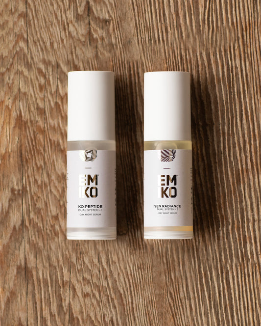 Two skincare serum bottles from the brand EMIKO, standing upright on a rustic wooden surface. The left bottle is labelled 'EMIKO KO PEPTIDE SERUM' and the right bottle is labelled 'EMIKO SEN RADIANCE SKIN SERUM'.