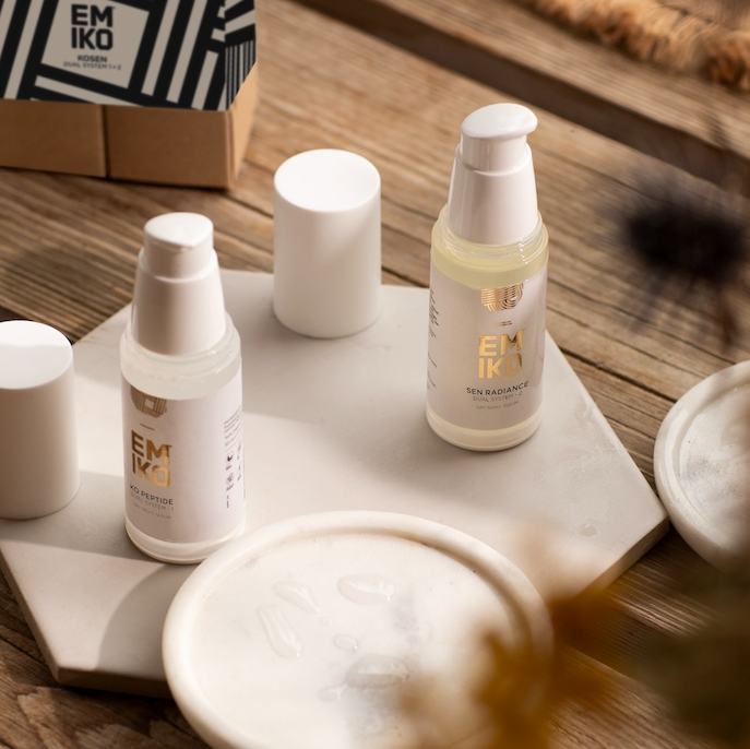 EMIKO SKINCARE, Two skincare bottles with air pump dispensers on a white stone tray on a rustic wooden table alongside skincare accessories. These are Kosen - patent pending dual system serum: ko: advanced peptides. Sen: bakuchiol, vitamin c thd ascorbate, vitamin e, seaweed 